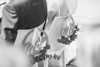 A black and white photograph showing the back view of a bride and groom seated on chairs decorated with circular signs reading 'Mr' and 'Mrs'. The chairs are adorned with foliage, and the bride is wearing a veil.