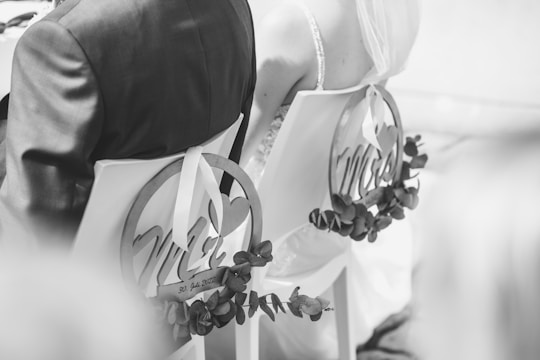 A black and white photograph showing the back view of a bride and groom seated on chairs decorated with circular signs reading 'Mr' and 'Mrs'. The chairs are adorned with foliage, and the bride is wearing a veil.