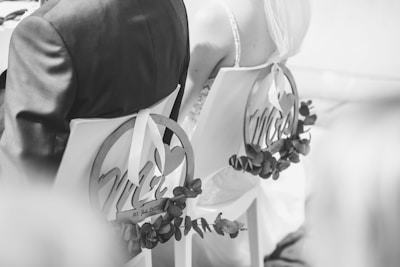 A black and white photograph showing the back view of a bride and groom seated on chairs decorated with circular signs reading 'Mr' and 'Mrs'. The chairs are adorned with foliage, and the bride is wearing a veil.