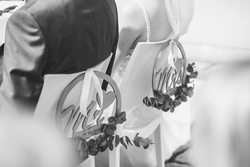 A black and white photograph showing the back view of a bride and groom seated on chairs decorated with circular signs reading 'Mr' and 'Mrs'. The chairs are adorned with foliage, and the bride is wearing a veil.