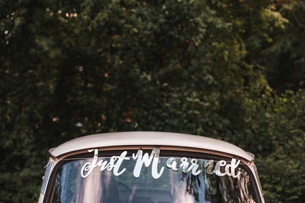 A vintage car with a 'Just Married' sign on the rear window is positioned in front of a lush, green forest background.