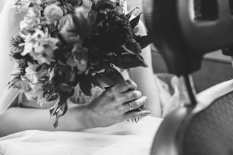 A monochrome photograph features a hand holding a bouquet of flowers. The focus is on the intricate details of the blossoms, arranged beautifully with various flowers and leaves. The hand is adorned with a ring, suggesting a special occasion. The setting appears to be indoors, possibly in a vehicle, as indicated by the car seat in the foreground.