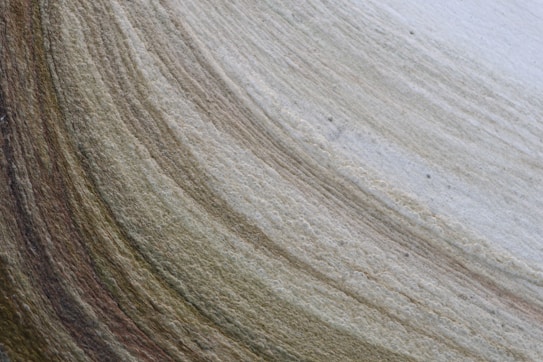 A close-up view of layered rock or sediment, showcasing intricate textures and stratified patterns. The layers vary in color, with subtle transitions from earthy browns to pale beiges and greys.