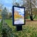 A digital billboard stands in a park displaying an autumn scene of a path lined with trees whose leaves are turning yellow. Surrounding the billboard, the park features green grass, a few scattered fallen leaves, trimmed hedges, and tall trees with orange and yellow foliage. The sky is partly cloudy, casting soft light over the area.