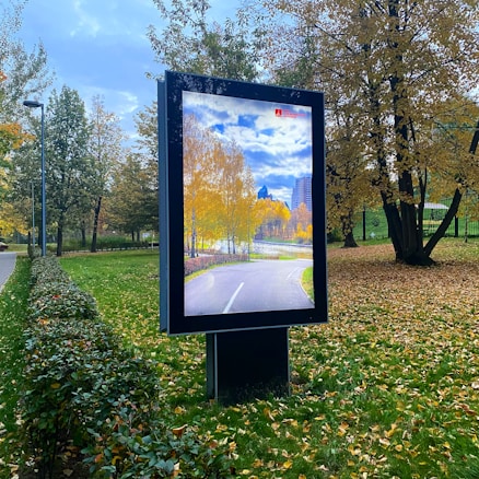 A digital billboard stands in a park displaying an autumn scene of a path lined with trees whose leaves are turning yellow. Surrounding the billboard, the park features green grass, a few scattered fallen leaves, trimmed hedges, and tall trees with orange and yellow foliage. The sky is partly cloudy, casting soft light over the area.