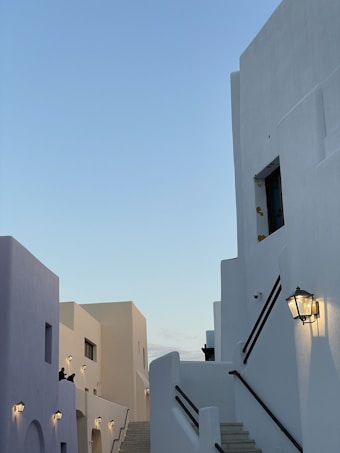 The image features white buildings with smooth, minimalist architecture and black railings, set against a clear blue sky. Soft lighting illuminates the walls, creating a tranquil and serene atmosphere. Outdoor lanterns are attached to the buildings, and a staircase is prominently featured.