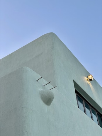 The image features a close-up view of the corner of a modern, minimalist building with smooth, light green stucco walls. A single outdoor wall light fixture is mounted near the top, illuminating the area softly. The sky is clear and blue, providing a stark contrast to the building's simple geometric lines. Three short, metallic pipes protrude from the wall, adding an industrial touch.