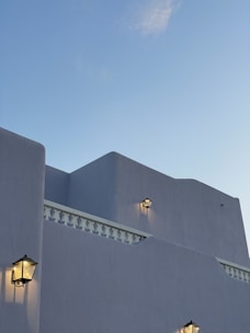 The image features a minimalist architectural structure with smooth, light gray walls and multiple lantern-style wall lights casting a warm glow. The building includes some intricate decorative railing. The sky in the background is clear with a hint of soft clouds.