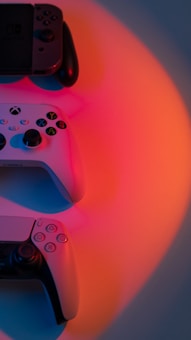 Three game controllers are arranged against a backdrop illuminated by vibrant red and orange lighting. Each controller has distinct buttons and joysticks, representing different gaming consoles.