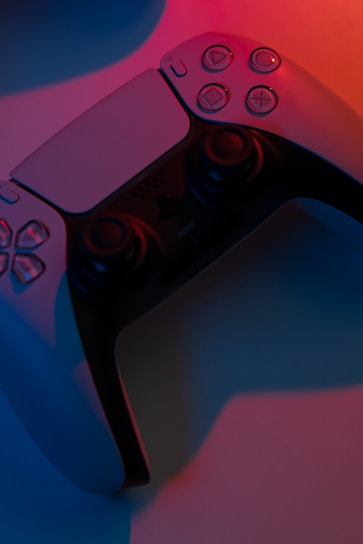 A close-up view of a game controller illuminated by contrasting blue and red lighting, providing a dramatic shadow effect. The controller features button details and analog sticks, reflecting modern gaming design aesthetics.