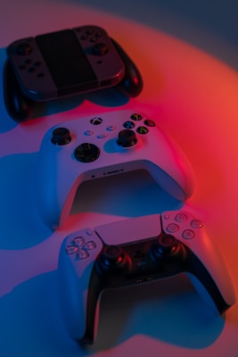 Three different gaming controllers are arranged on a surface illuminated by blue and red lighting. The controllers have distinct designs, featuring various buttons and control sticks.