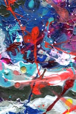 A colorful abstract painting with vibrant splashes and swirls of red, blue, pink, white, and green. The textures are dynamic, with layers overlapping and mixing in a chaotic yet harmonious manner. The paint appears to be thickly applied, creating a tactile surface.