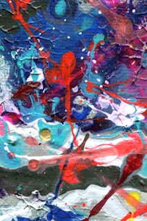 A colorful abstract painting with vibrant splashes and swirls of red, blue, pink, white, and green. The textures are dynamic, with layers overlapping and mixing in a chaotic yet harmonious manner. The paint appears to be thickly applied, creating a tactile surface.