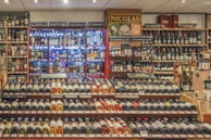 A well-stocked wine and spirits store with numerous bottles of wine displayed on wooden shelves. The shelves feature a range of wines from different regions and spirits in glass bottles, illuminated by soft lighting. Signs indicate different categories such as liqueurs, wines, and special beers.
