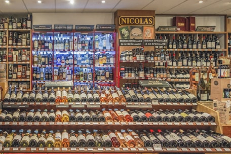 A well-stocked wine and spirits store with numerous bottles of wine displayed on wooden shelves. The shelves feature a range of wines from different regions and spirits in glass bottles, illuminated by soft lighting. Signs indicate different categories such as liqueurs, wines, and special beers.