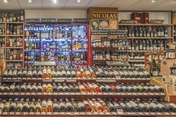 A well-stocked wine and spirits store with numerous bottles of wine displayed on wooden shelves. The shelves feature a range of wines from different regions and spirits in glass bottles, illuminated by soft lighting. Signs indicate different categories such as liqueurs, wines, and special beers.