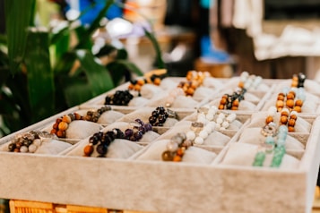 A display of beaded bracelets presented on a soft, cushioned tray. The assortment includes various colors and textures, such as white, black, brown, and green beads situated in individual compartments. The background shows some greenery, adding a natural element.
