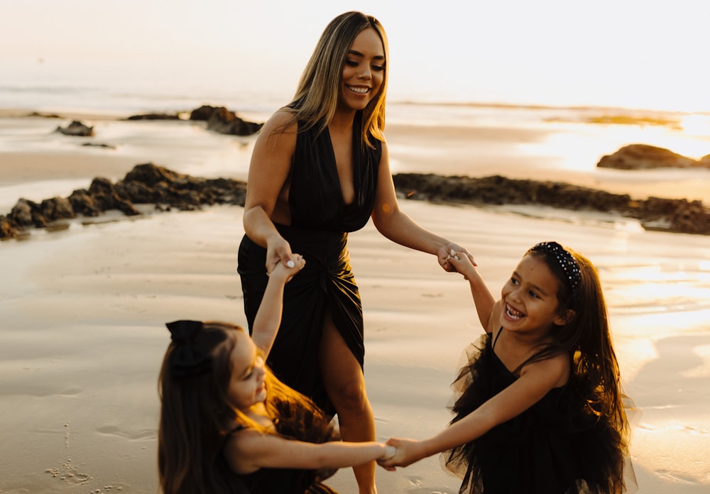 a woman in a black dress holding hands with two girls on the beach
