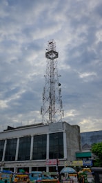 A tall communication tower stands against a cloudy sky, positioned above a building with dark glass windows. Below the tower, there are colorful street vendors and food stalls lined up, adding vibrancy to the scene.