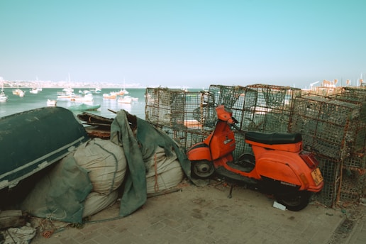 A red scooter is parked near a stack of fishing cages or lobster pots by the seaside. In the background, several small boats are floating on calm water under a clear blue sky. The scene gives a rustic, coastal vibe with industrial elements.