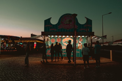 An outdoor food stall is illuminated during early evening, featuring a turquoise and pink theme. Several silhouettes of people are gathered in front of the stall, which offers churros, farturas, and waffles. The background includes a dimly lit, colorful amusement ride and a clear sky transitioning from orange to teal.