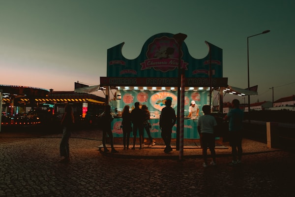 An outdoor food stall is illuminated during early evening, featuring a turquoise and pink theme. Several silhouettes of people are gathered in front of the stall, which offers churros, farturas, and waffles. The background includes a dimly lit, colorful amusement ride and a clear sky transitioning from orange to teal.