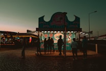 An outdoor food stall is illuminated during early evening, featuring a turquoise and pink theme. Several silhouettes of people are gathered in front of the stall, which offers churros, farturas, and waffles. The background includes a dimly lit, colorful amusement ride and a clear sky transitioning from orange to teal.