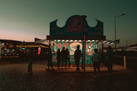 An outdoor food stall is illuminated during early evening, featuring a turquoise and pink theme. Several silhouettes of people are gathered in front of the stall, which offers churros, farturas, and waffles. The background includes a dimly lit, colorful amusement ride and a clear sky transitioning from orange to teal.