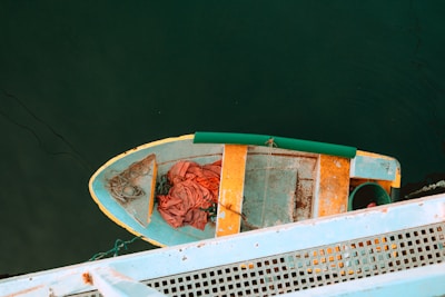 An aerial view of a small, weathered boat with vibrant colors, floating on dark water. The boat is painted in shades of yellow, blue, and orange, with some wear visible. A green cylindrical object is placed on the dock beside the boat, and there is a tangled rope and fabric inside the boat. The dock has a grid texture and is painted light blue.