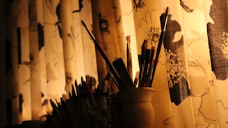 A collection of paintbrushes is arranged in a container against a backdrop of floral-patterned curtains. The warm lighting casts soft shadows, creating an inviting and serene atmosphere.