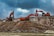 Two large excavators engage in construction work on a rocky site, with overcast clouds adding a dramatic backdrop. The machinery, robust in design and red in color, dominates the scene amidst the earthy tones of the rocky terrain.