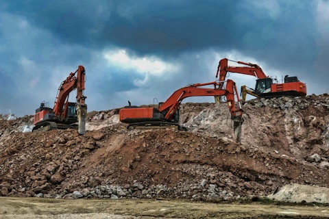 Two large excavators engage in construction work on a rocky site, with overcast clouds adding a dramatic backdrop. The machinery, robust in design and red in color, dominates the scene amidst the earthy tones of the rocky terrain.