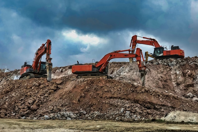 Two large excavators engage in construction work on a rocky site, with overcast clouds adding a dramatic backdrop. The machinery, robust in design and red in color, dominates the scene amidst the earthy tones of the rocky terrain.