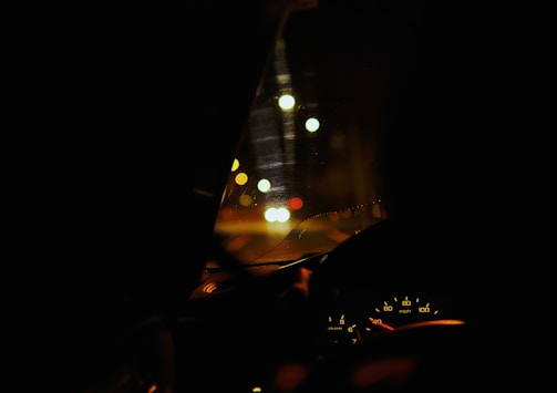 A dimly lit car interior with a focus on the dashboard and the speedometer, showing a speed of around 40 mph. The view through the windshield features bokeh effects from street lights or traffic lights, creating a blurred background with a mix of warm colors.