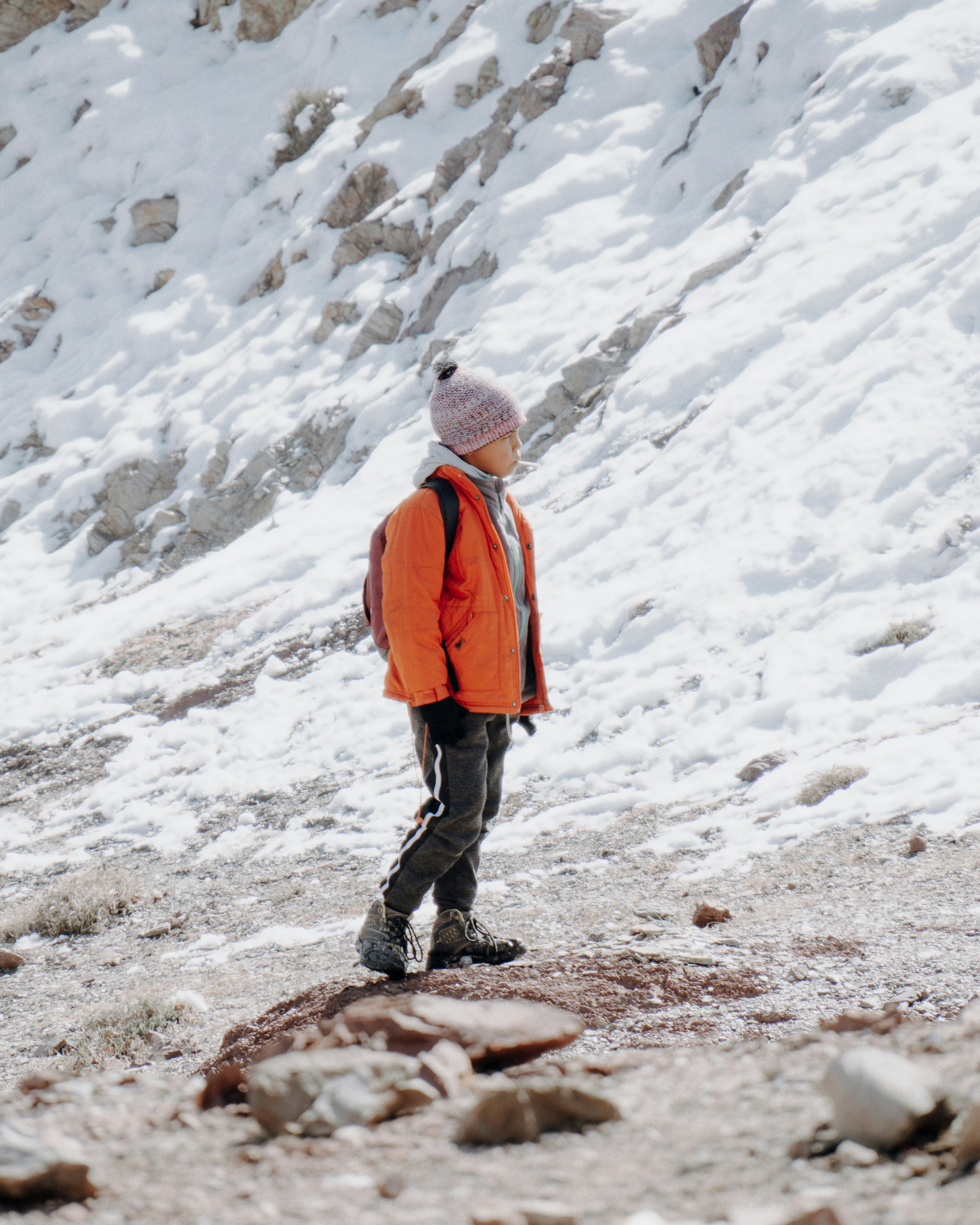 Young hiker in an orange insulated jacket, knit hat and small pack walking across a snowy slope with rocks showing through