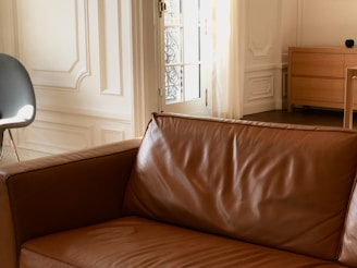 A well-lit interior space featuring a brown leather sofa in the foreground. In the background, there is a white paneled wall and a partially visible modern chair with a grey cushion. Natural light streams in through a door with an ornate metal grille. A light wooden chest of drawers is positioned to the right.