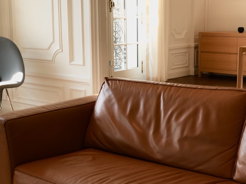 A well-lit interior space featuring a brown leather sofa in the foreground. In the background, there is a white paneled wall and a partially visible modern chair with a grey cushion. Natural light streams in through a door with an ornate metal grille. A light wooden chest of drawers is positioned to the right.