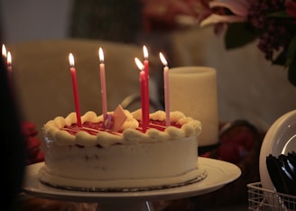 A round cake with white frosting and red jelly topping decorated with lit pink candles. There is a small pink and white striped decoration on top of the cake. To the right, plastic cutlery is arranged in a holder, and a large candle is unlit in the background.