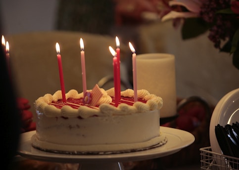 A round cake with white frosting and red jelly topping decorated with lit pink candles. There is a small pink and white striped decoration on top of the cake. To the right, plastic cutlery is arranged in a holder, and a large candle is unlit in the background.