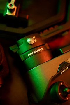 A close-up of a mechanical device labeled 'PAN AXIS MOTOR' with intricate details. The image is illuminated with green and red lighting, creating a striking contrast. Metallic components and switches are visible, suggesting technological and industrial elements.