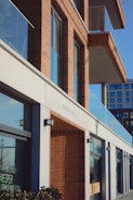 A modern building with a facade of red brick and glass, featuring multiple balconies and large windows. The lower part of the building has storefronts with signage visible through the windows. The architecture is contemporary, with clean lines and a combination of materials including concrete, glass, and brick.