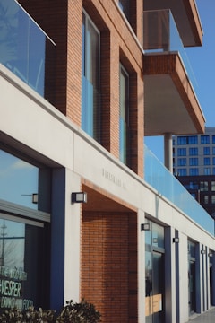 A modern building with a facade of red brick and glass, featuring multiple balconies and large windows. The lower part of the building has storefronts with signage visible through the windows. The architecture is contemporary, with clean lines and a combination of materials including concrete, glass, and brick.