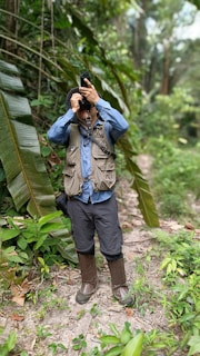 A person standing on a dirt trail in a dense forest. They are equipped with hiking gear, including a vest with multiple pockets, long pants, gaiters, and a hat. The individual is holding a camera up to their face, seemingly taking a photograph. The surrounding area is lush and green with various types of vegetation, including large banana leaves.