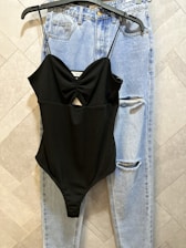A pair of light blue denim jeans with a ripped detail near the knee is hanging on a tile-patterned wall. Over the jeans hangs a black spaghetti-strap bodysuit with a central cutout design.