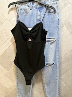 A pair of light blue denim jeans with a ripped detail near the knee is hanging on a tile-patterned wall. Over the jeans hangs a black spaghetti-strap bodysuit with a central cutout design.