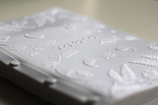A close-up view of a book or booklet with pages that have raised designs and patterns embossed onto them. The surface features intricate floral motifs, small shapes, and lines, suggesting a tactile element to the material.