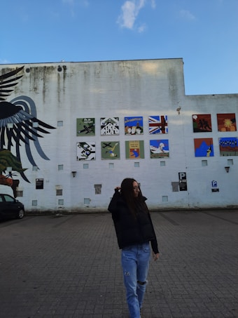 A large white wall with various colorful murals is prominently featured. A stylized black bird mural stretches across one side, and multiple square paintings depicting different scenes are aligned in a grid. The sky is clear with a few clouds. In the foreground, a person wearing a black jacket and ripped jeans walks in front of the wall, with a parking area visible beneath them.