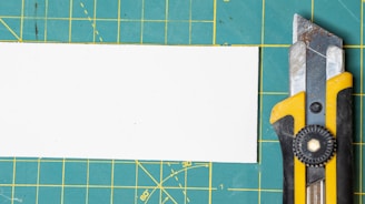 A utility knife with a yellow and black handle is placed on a green cutting mat with a grid pattern. There is a white rectangular piece of paper or board beside the knife.