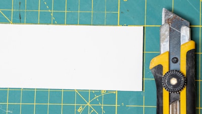 A utility knife with a yellow and black handle is placed on a green cutting mat with a grid pattern. There is a white rectangular piece of paper or board beside the knife.