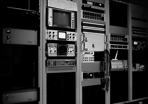 A collection of vintage electronic equipment is mounted in tall racks, featuring a variety of dials, knobs, and screens. Each unit appears to be a specialized piece of technology, possibly related to broadcasting or audio engineering. Cables are visibly hanging down the side of one rack.